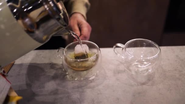キッチンテーブルで熱いお茶を作っています 沸騰した水はケトルからティーバッグ付きのカップに注ぎ込まれます キッチンテーブルに新鮮なお茶の透明なカップ — ストック動画