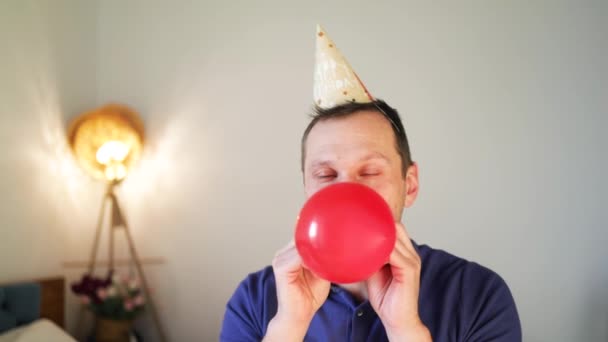 Jonge Blanke Man Met Een Verjaardagspet Die Rode Ballon Opblaast — Stockvideo
