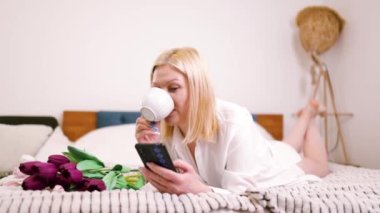Güzel beyaz kadın yatak odasındaki yatakta uzanıyor. Akıllı telefonu çevrimiçi alışveriş için beklet. Çiçekler yakında..