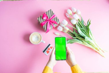 Kadın elinin yeşil renkli ekranlı cep telefonuyla bir fincan kahve, kredi kartı ve çiçeklerle çekilmiş en iyi görüntüsü.