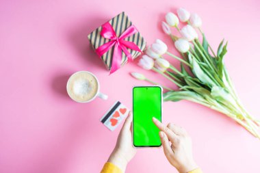 Kadın elinin yeşil renkli ekranlı cep telefonuyla bir fincan kahve, kredi kartı ve çiçeklerle çekilmiş en iyi görüntüsü.