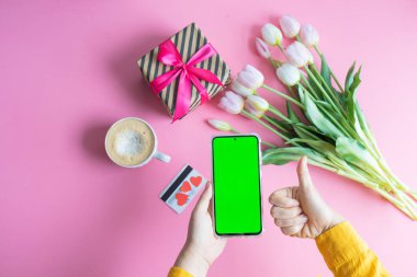 Kadın elinin yeşil renkli ekranlı cep telefonuyla bir fincan kahve, kredi kartı ve çiçeklerle çekilmiş en iyi görüntüsü.