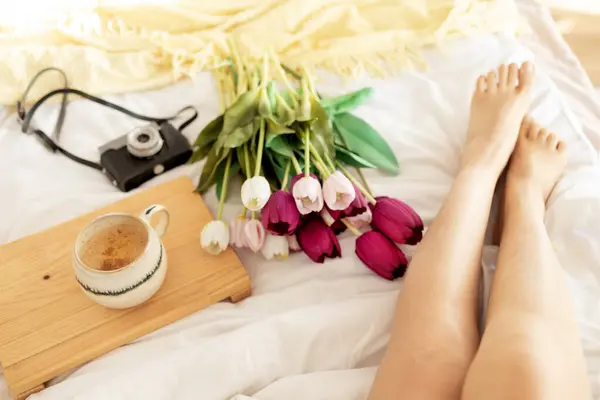 Female feet with bouquet of tulips and coffee on bed.