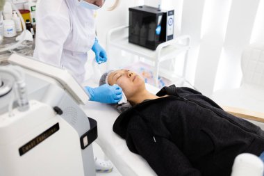 Kozmetik uzmanı, kozmetik cerrahi kliniğinde yüz mezoterapisini temizlerken hastanın yanaklarına krem sürüyor..