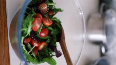 Mutfak konsepti, ev yapımı yemekler için hazırlama malzemeleri. Dikey çekim, yemek pişiren tanınmayan bir kadını vurdu. Salatayı içeride salata kasesine döktü. Sebzeler masada.