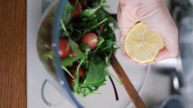 Mutfak konsepti, ev yapımı yemekler için hazırlama malzemeleri. Dikey çekim, tanınmayan bir kadın mutfakta limon suyuyla salata pişiriyor. Sebzeler masada..