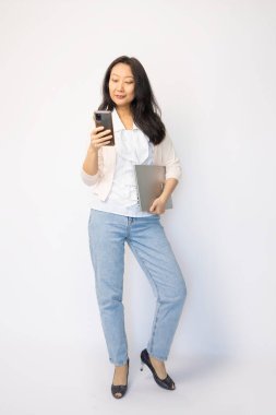 Sırıtkan Asyalı bir iş kadınının tam boy portresi dizüstü bilgisayar ve akıllı telefon taşırken beyaz arka planda tek başına duruyor.