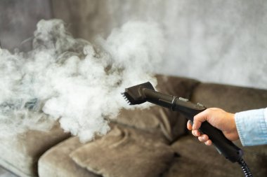 Ev temizliği. Bir adam kanepeyi ve mobilyaları buhar jeneratörüyle temizliyor. Dairenin temizlik servisi. Düz Los. Güçlü buhar basıncı.