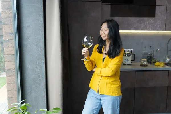 Waist up of relaxed young Asian woman in yellow sweater smiling and looking at the glass of wine in her hand while standing in the kitchen.