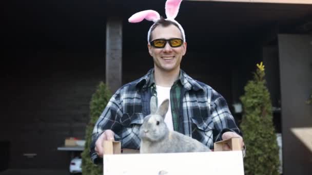 照片中快乐而又滑稽的英俊年轻人站在家门口 目不转睛地看着相机 抱着复活节兔子 微笑着 — 图库视频影像