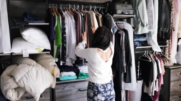 若いアジアの女性は 家や店のクローゼットリビングルームで彼女のファッション服を選択します 女の子はカジュアルなシャツを着るために何を着るべきか考えます ビッグホームワードローブ — ストック動画