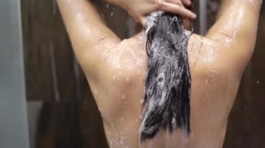 Arka planda, uzun siyah saçlı genç bir Asyalı kadının modern bir banyoda duşun altında durup saçlarını yıkaması ve durulaması var..