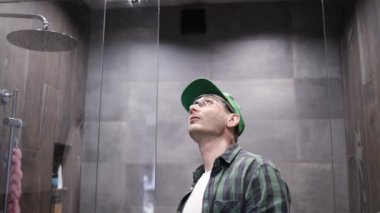 Üniformalı genç adam banyodaki duş kapısını tamir ediyor. Erkek bir tamirci duş kabinini tamir ediyor..