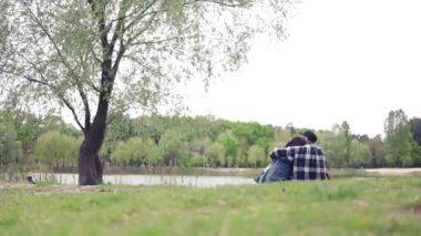 Genç çift göl kenarındaki çimlerin üzerinde oturup birbirlerine sarılıyorlar. Güzelim bahar gününde çimlerin üzerinde oturan genç çiftin arka görünüşü.
