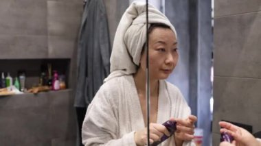 Orta yaşlı, kafasında bağırsak olan Asyalı bir kadın, banyoda sabah duşundan sonra nemlendirici krem sürüyor. Mutlu Çinli kadın cildini suluyor, günlük cilt bakımının keyfini çıkarıyor..