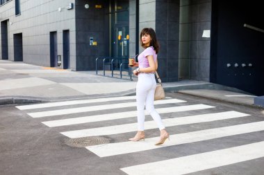 Beyaz kotlu bej çantalı ve topuklu ayakkabılı genç bir kadının modern binanın yakınındaki sokakta gezinip kahve içerkenki görüntüsü..