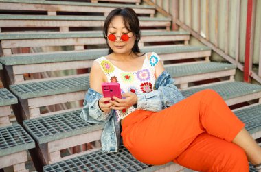 Moda Asyalı bir kadının yaz günü sokak merdivenlerinde oturup şehirdeki cep telefonuna bakması..