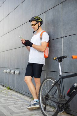 Şehirde telefonunu kontrol eden formda, mutlu bir bisikletçi. Rahatlamış gülümseyen atletik bisikletli erkek akıllı telefonunu duvara yaslanmış izliyor..