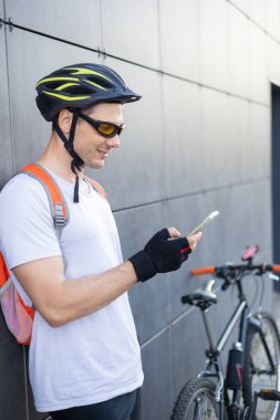 Şehirde telefonunu kontrol eden formda, mutlu bir bisikletçi. Rahatlamış gülümseyen atletik bisikletli erkek akıllı telefonunu duvara yaslanmış izliyor..