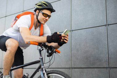 Şehirde telefonunu kontrol eden formda, mutlu bir bisikletçi. Rahatlamış gülümseyen atletik bisikletli erkek akıllı telefonunu duvara yaslanmış izliyor..