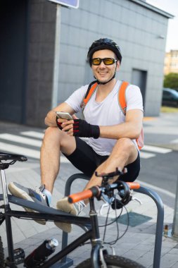 Bisikletli genç adam yaya geçidinin yanında telefonunu kontrol ediyor. Rahatlamış gülümseyen atletik bisikletli erkek akıllı telefonunu şehrin duvarında dinlenirken izliyor..