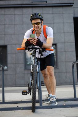 Bisikletli genç adam yaya geçidinin yanında telefonunu kontrol ediyor. Rahatlamış gülümseyen atletik bisikletli erkek akıllı telefonunu şehrin duvarında dinlenirken izliyor..