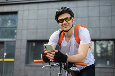 Bisikletli genç adam yaya geçidinin yanında telefonunu kontrol ediyor. Rahatlamış gülümseyen atletik bisikletli erkek akıllı telefonunu şehrin duvarında dinlenirken izliyor..
