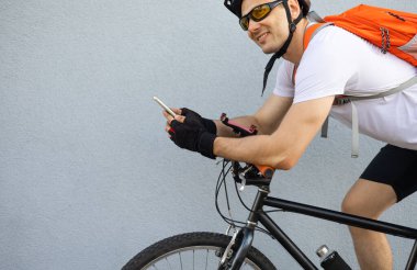Şehirde telefonunu kontrol eden formda, mutlu bir bisikletçi. Rahatlamış gülümseyen atletik bisikletli erkek akıllı telefonunu duvara yaslanmış izliyor..