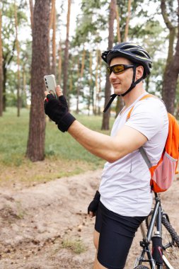 Yakışıklı beyaz bisikletçi yaz ormanında akıllı telefonuyla görüntülü konuşma yapıyor. Genç sporcu bisikletinin yanında selfie çekiyor.. 