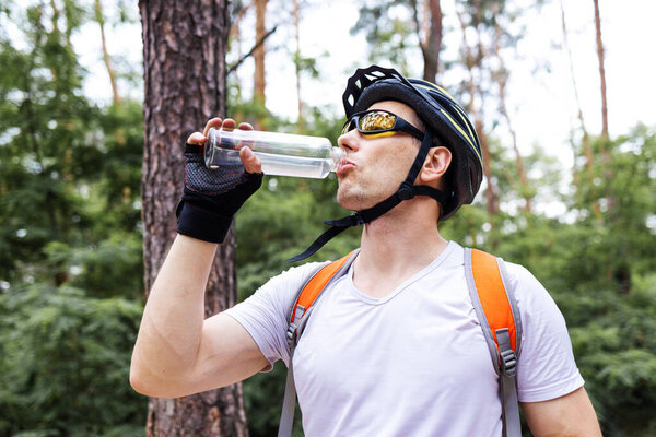 Жаждущий профессиональный велосипедист держит бутылку с водой, стоя на велосипеде в парке в солнечный день.