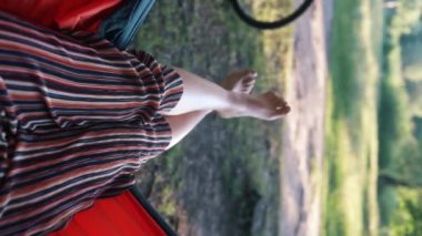 Ormanda güneşli bir yaz gününde hamakta bir kadının bacaklarına bakış açısı. Dikey video.
