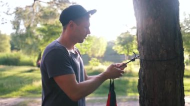 Erkek turist setleri kampa bisikletle geldi. Bisikletçi ormanda hamak kuruyor. Gezgin parkta hamak bağlıyor. Adam ağaca hamak kayışı takıyor..