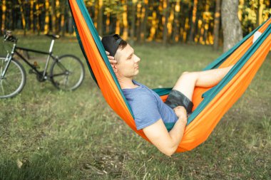 Genç adam bisiklete biniyor, kırmızı hamakta dinleniyor, gölün yakınındaki ormanda dinleniyor. Hamakta nehir kenarındaki kamp alanında mutlu bisikletçiler. Günbatımında hamakta bisikletli bir erkek.