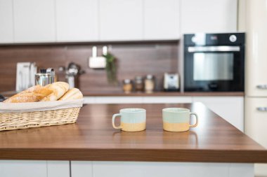 Mutfaktaki ahşap masanın üzerinde beyaz dolaplar, kahve fincanları ve kruvasanlar olan minimalist bir tasarım sergiliyor..