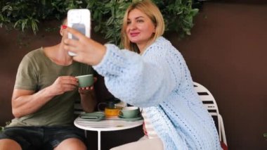 Güneşli bir sonbahar gününde açık havada oturan genç çift. Teras Kafe 'de sipariş bekliyor, akıllı telefon bakıyor, selfie çekiyor..