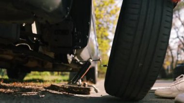 Arka plandaki genç adam arabasının lastiğini değiştirdi. Tekerlek değiştiren bir tamircinin son sahnesi. Arabanın tekerleği değişiyor, yol kenarında yedek lastik var.