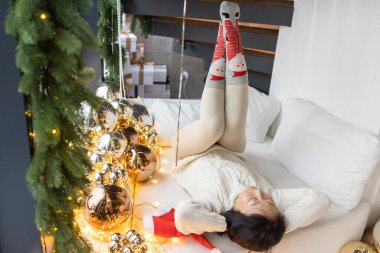 Genç Asyalı kadın Noel sabahı pencerenin yanında oturuyor. Bir kış günü apartmanının sıcacık ortamında şenlik kıyafeti ve kırmızı Noel çorapları giyen bir kadın.