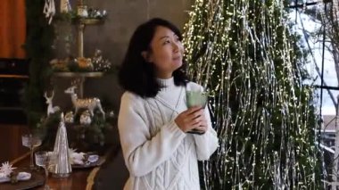 Mutlu Asyalı kadın elinde şampanya bardağıyla Noel ağacının önünde hayal kuruyor.