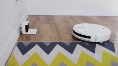 Beyaz robotik elektrikli süpürge temizliği bitirir ve şarj istasyonuna gider. Robot elektrikli süpürge odadaki zemini temizliyor. Ev kadınlarının temizlik yapmasına yardımcı olan modern teknolojiler.