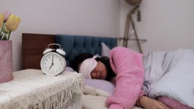 Uykulu Asyalı kız battaniyeye sarılıp yavaş çekimde uyumaya devam ediyor. Yatakta uyuyan Asyalı bir kadın sabahın erken saatlerinde komodindeki dijital çalar saati etkisiz hale getirmek için uyanıyor..