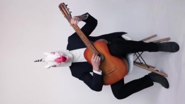 Takım elbiseli, at kafası atlayan ve beyaz arka planda gitar çalan komik bir genç. Dikey video.