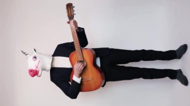 Takım elbiseli, at kafası atlayan ve beyaz arka planda gitar çalan komik bir genç. Dikey video.