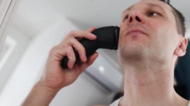 Beyaz tişörtlü beyaz bir adam evinde tıraş olurken elektrikli jiletle tıraş oluyor..