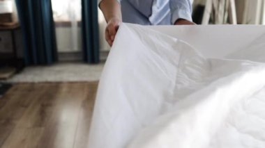 Kadın kapalı bir yerde şiltenin üstüne örtü örttü, yakın plan. Yatağın şiltesine elastik bantlı beyaz bir saten çarşaf konur, ellerin yakın çekimi. Yatak örtüleri.