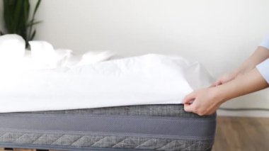 Kadın kapalı bir yerde şiltenin üstüne örtü örttü, yakın plan. Yatağın şiltesine elastik bantlı beyaz bir saten çarşaf konur, ellerin yakın çekimi. Yatak örtüleri.