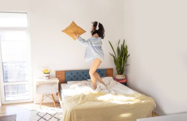 Uzun boylu, kulaklıklı, neşeli Asyalı bir kadının evde yatakta şarkı söyleyip zıplaması..