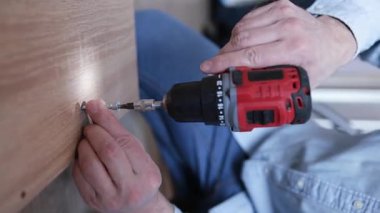 Bir adam elektrikli matkapla dolaba vida sokuyor. Mobilya montajı. Dikey video.