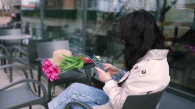 Mesajlaşma, iletişim ve içerik için telefonunu kullanan şık Asyalı kadın açık hava kafede oturan sosyal medya için yaratıyor. Lale buketi tutan kız. Etkileyici..