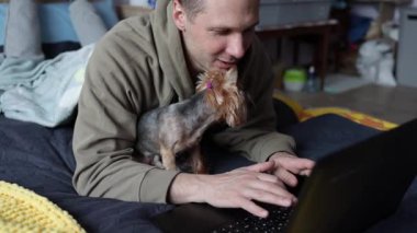 Genç adam evde yatakta otururken dizüstü bilgisayar kullanıyor ve köpeğe dokunuyor. Beyaz adamın yakın görüntüsü görüntüye bakıyor ve uzaktan iş yapıyor, sevimli hayvanlara dokunuyor ve iç kısımda yatağın yanında oturuyor.. 