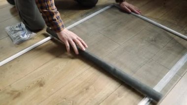 Pencere örgü ören ellere yakın çekim. Parmaklar ağ ile çerçeve kanalına basar. Bulanık arka plan, kapalı mekan ayarlarını gösteriyor. Ekran malzemelerinin korunması üzerine titiz ve detaylı çalışmalara odaklanın